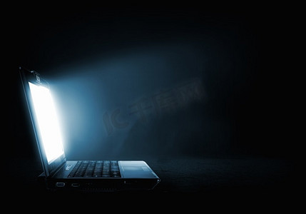暗背景摄影照片_发光的笔记本电脑。打开笔记本电脑，光线来自屏幕，背景为暗
