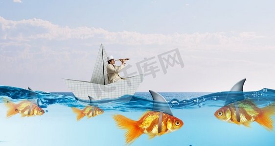 漂浮金鱼摄影照片_给您的业务带来虚假风险。假威胁的概念当商人漂浮在纸船上，鲨鱼在水中看起来像金鱼