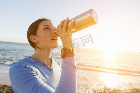 一名妇女在海滩上喝水。年轻女子在海滩上运动后喝水