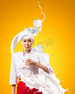 拿牛奶摄影照片_亚洲女厨师对牛奶飞溅。亚洲女性厨师反对牛奶飞溅在红色围裙反对拿着牛奶玻璃的颜色背景