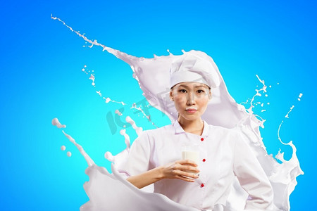 亚洲女厨师对牛奶飞溅。亚洲女性厨师反对牛奶飞溅在红色围裙反对拿着牛奶玻璃的颜色背景