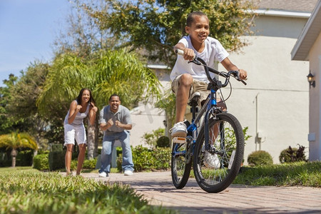 一个年轻的非洲裔美国家庭，男孩骑着自行车，他的快乐兴奋的父母在他身后给予鼓励。