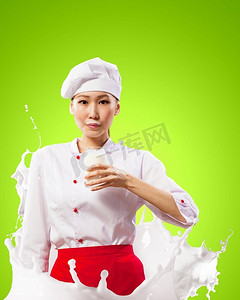 亚洲女厨师防止牛奶溅起。亚洲女厨师手持一杯牛奶，穿着红色围裙，手持牛奶，在彩色背景下飞溅