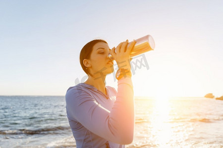 出水素果摄影照片_一名妇女在海滩上喝水。年轻女子在海滩上运动后喝水