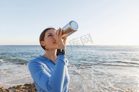 一名妇女在海滩上喝水。年轻女子在海滩上运动后喝水