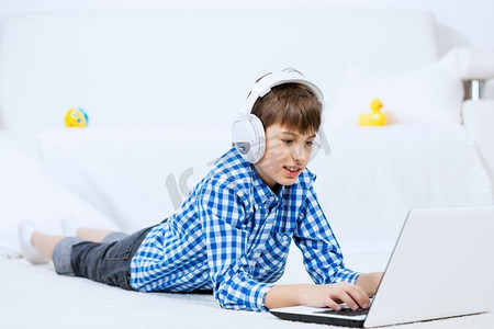 音乐歌手刘德华摄影照片_享受休闲时光的孩子。学龄儿童躺在地上通过耳机听笔记本电脑上的音乐