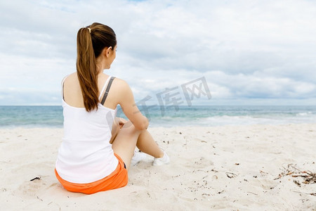 一分钟摄影照片_一位穿着运动服的年轻女子坐在海滩上。穿着运动服的年轻女子独自坐在海滩上休息一分钟