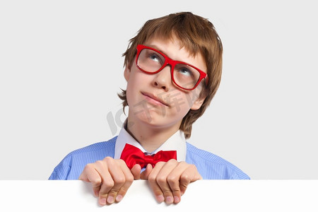 红白色海报摄影照片_戴红眼镜的男孩拿着白色方块。小男孩若有所思地微笑着拿着白色方块的形象。刊登广告的地点