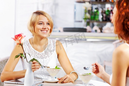 咖啡厅女孩摄影照片_咖啡馆里的可爱女孩。两个年轻漂亮的女人坐在咖啡馆里吃着甜点