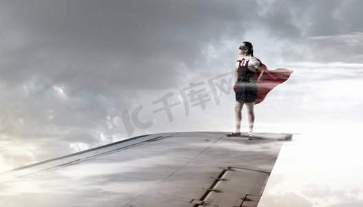 小超级英雄。强大的超级英雄小女孩站在飞行的飞机机翼上