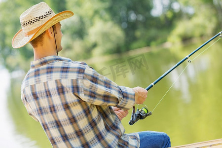 夏季钓鱼。戴着帽子的年轻人坐在桥上钓鱼