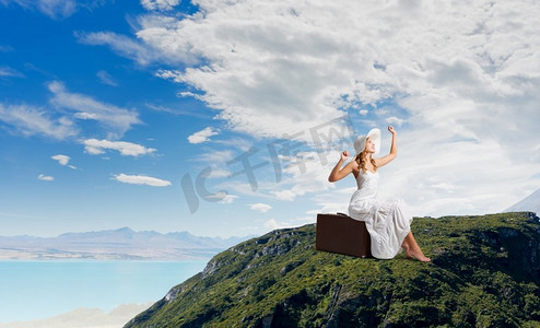 她轻装上阵。一位穿着白色长裙、戴着帽子的女子坐在山顶的行李上