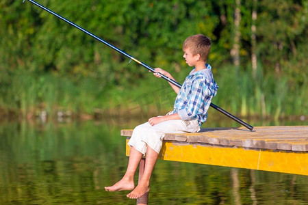 穿着蓝色衬衫的男孩坐在海边的码头上，手里拿着钓鱼竿