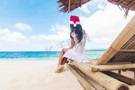 圣诞老人帽子摄影照片_一名妇女在海滩上庆祝圣诞节。戴着圣诞老人帽子的年轻女子坐在热带海滩的沙发休息室里庆祝圣诞节