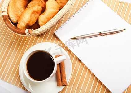 金色钢笔背景摄影照片_在浅色背景下提供早餐咖啡和牛角面包，桌上有笔记本记录。