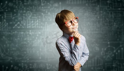 黑板公式摄影照片_聪明的小学生。戴着红眼镜的天才男孩在黑板上写着公式
