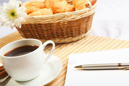 在浅色背景下提供早餐咖啡和牛角面包，桌上有笔记本记录。