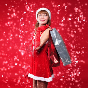 购物袋的女孩摄影照片_红色背景上，一位打扮成圣诞老人的年轻女孩正在购物。一个拿着购物袋的女孩。双旦快乐!