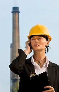一位美丽的黑发女子，戴着安全帽，站在工厂及其烟囱前打着手机