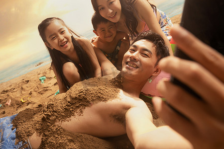 泳衣男人摄影照片_沙滩上男人拿手机和妻子孩子自拍