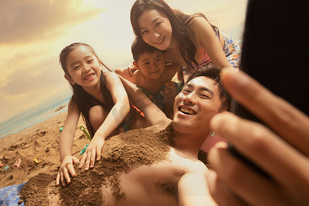 沙滩上男人拿手机和妻子孩子自拍
