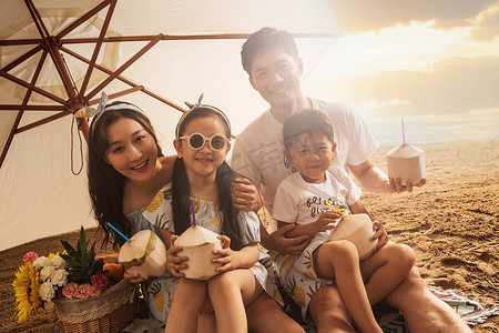 夏天饮料吸管摄影照片_幸福的四口之家坐在沙滩上喝椰汁