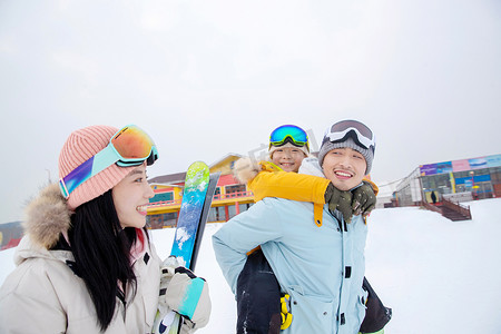 雪地玩耍人物摄影照片_滑雪场上背着儿子的父亲和抱着滑雪板的母亲