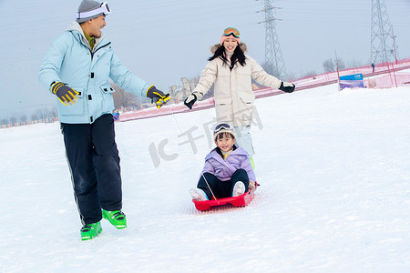 雪地玩耍人物摄影照片_滑雪场上父母和坐在雪上滑板的女儿玩耍