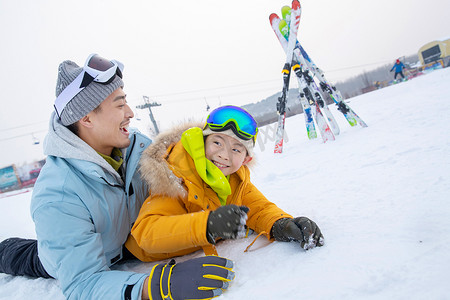 雪地玩耍人物摄影照片_滑雪场内抱在一起打滚的快乐父子