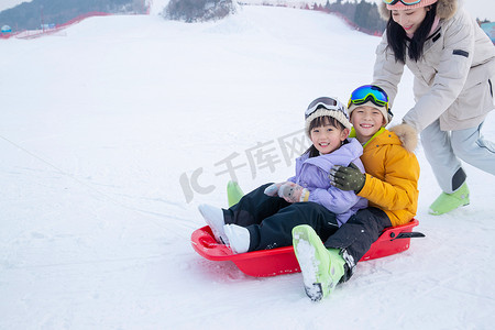 雪地玩耍人物摄影照片_雪场上妈妈推着坐在雪上滑板的孩子们