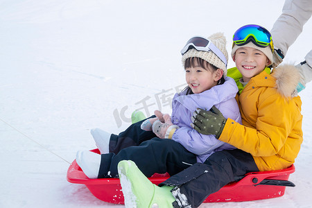 雪地玩耍人物摄影照片_雪场上推着坐在雪上滑板的孩子们