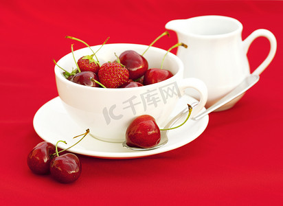 越南河粉摄影照片_红色背景下的牛奶壶杯子茶碟勺子樱桃和草莓