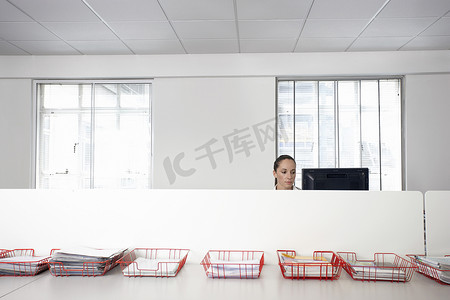 在装有文件的托盘后面的办公室隔间里使用电脑的女商人