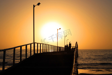 太阳橙色摄影照片_沿着拉格湾码头行走的人们身后的橙色日落，阿德尔
