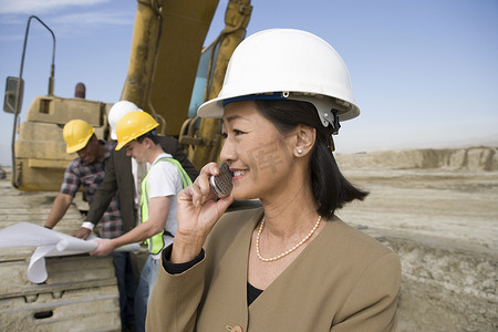 在现场使用手机的工人和重型机械前戴安全帽的女测量员