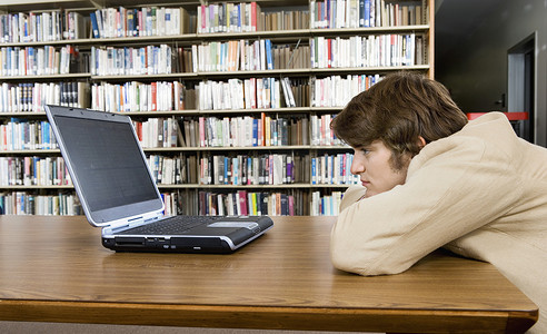 坐在图书馆办公桌前看着笔记本电脑的无聊男大学生的侧视图