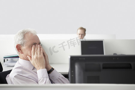 中年商人在电脑桌前与背景中的同事揉眼睛