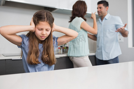 抑郁的孩子摄影照片_伤心的女孩捂着耳朵，父母在厨房里吵架