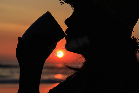 剪影咖啡杯摄影照片_泰国妇女在海滩上拿着咖啡杯喝酒的剪影
