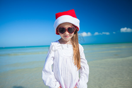 红色帽子女孩摄影照片_红色帽子圣诞老人和太阳镜的小女孩在异乎寻常的海滩