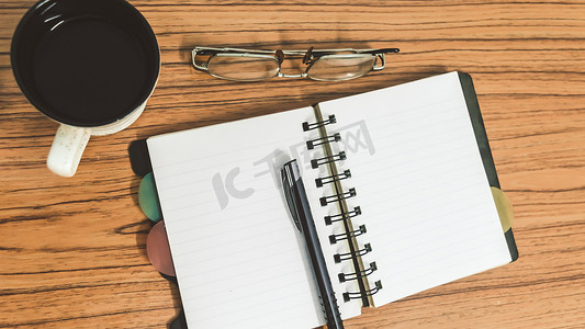 桌子上有打开的笔记本，上面有空白页、眼镜、笔和一杯咖啡。