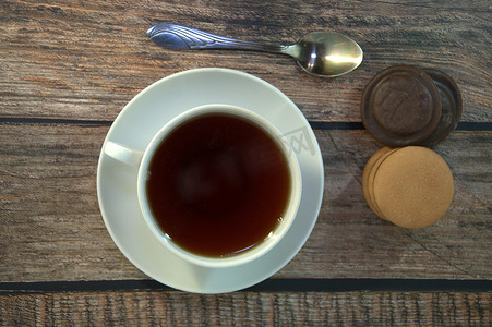 瓷碟摄影照片_木桌上放着一杯放在瓷碟上的红茶、一把勺子和一堆巧克力海绵饼干。