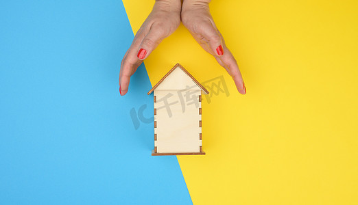 两只女性的手在黄蓝色背景的木制微型模型房子上相互交叉。