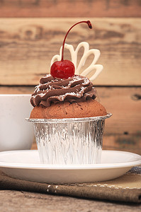 巧克力奶油杯子摄影照片_旧木背景中带巧克力奶油和樱桃的纸杯蛋糕