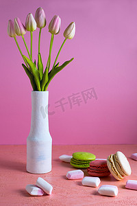 马卡龙和小的白色和粉色棉花糖散落在淡粉色背景上，旁边是一瓶郁金香。
