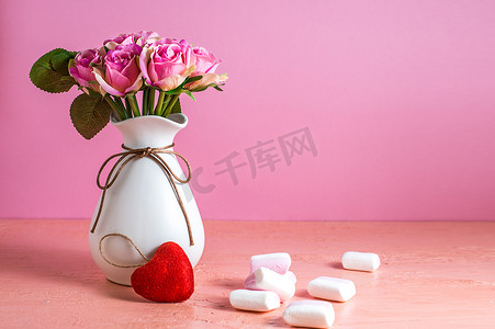 背景淡粉摄影照片_白色和粉色的小棉花糖散落在淡粉色的背景上，旁边是一瓶玫瑰。