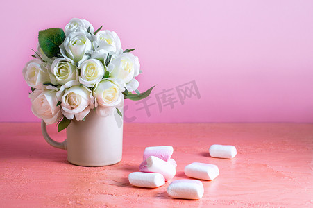 淡彩摄影照片_白色和粉色的小棉花糖散落在淡粉色的背景上，旁边是一瓶玫瑰。