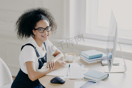 积极的非裔美国女性自由职业者在工作场所摆出论文和教科书的姿势，在电脑上远程工作，喝咖啡休息，在自己的橱柜里工作，穿着休闲服。