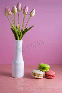淡彩摄影照片_淡粉色背景上的蛋白杏仁饼干，旁边是一瓶郁金香。