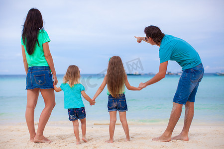 阳光明媚的衣服摄影照片_阳光明媚的日子里，在异国情调的海滩上，带着两个女儿的年轻家庭的背影